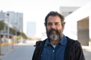 A mature white man stands on a city street in daytime. He has a tanned complexion and dark/grey beard. He is wearing a dark jacket and blue shirt with an open collar with a white t-shirt underneath. He is looking at the camera with a neutral expression.