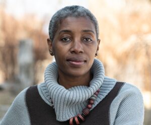 A middle aged Black woman sits outside against a city landscape. She has cropped dark and grey hair. She is wearing a brown and light grey shawl and a chunky necklace. She is looking into the camera with a neutral expression.