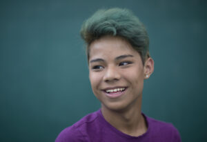 Portrait of a young Asian trans/non-binary person against a blue-grey backdrop. They are wearing a purple t-shirt and have pierced ears. They have short hair which is dyed blue at the tips. They are smiling and looking past the camera.
