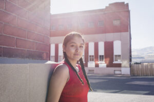 A Young Indigenous woman stands outside in a rural town. She is standing against the corner of a red brick building. Behind her you can see a wide street, more buildings, and hills in the distance. The sun is shining above her head. She has long dark hair in braids and is wearing a red/dark orange tank. She is looking at the camera with a friendly expression.